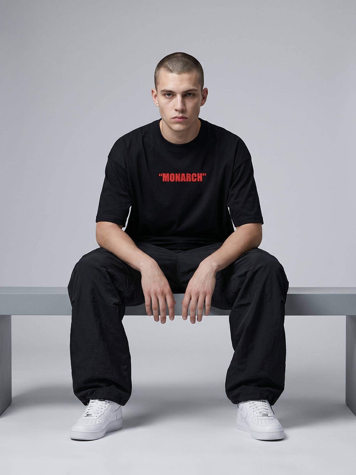 Person wearing a black t-shirt with 'MONARCH' printed in red, sitting on a bench against a gray background.