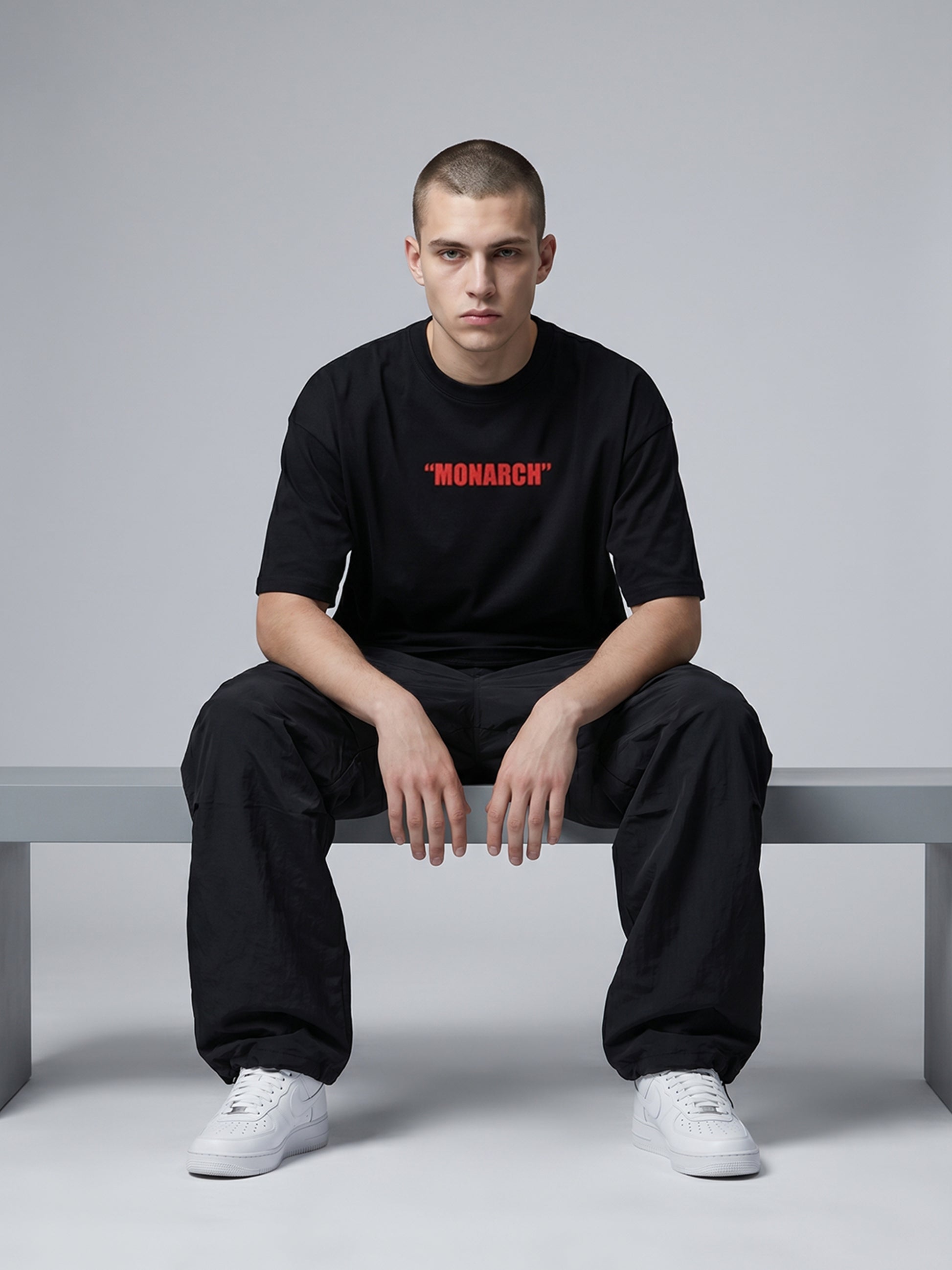 Person wearing a black t-shirt with 'MONARCH' printed in red, sitting on a bench against a gray background.