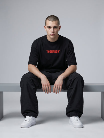 Person wearing a black t-shirt with 'MONARCH' printed in red, sitting on a bench against a gray background.