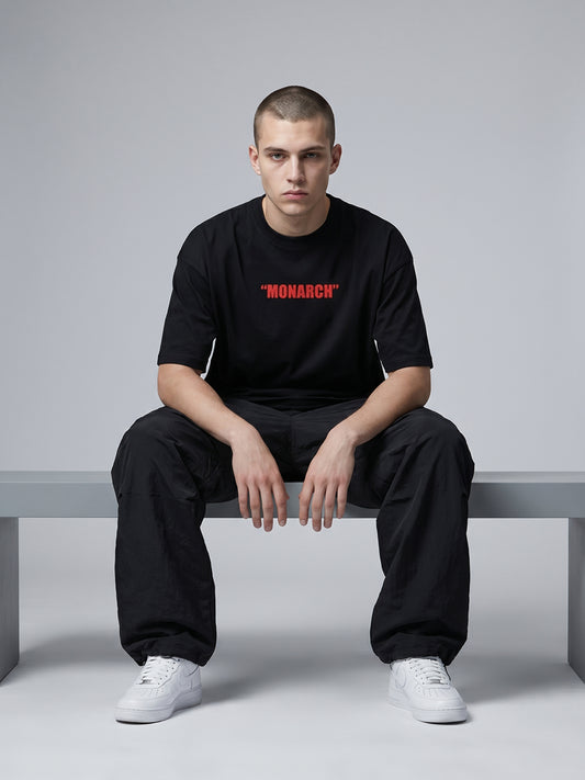 Person wearing a black t-shirt with 'MONARCH' printed in red, sitting on a bench against a gray background.