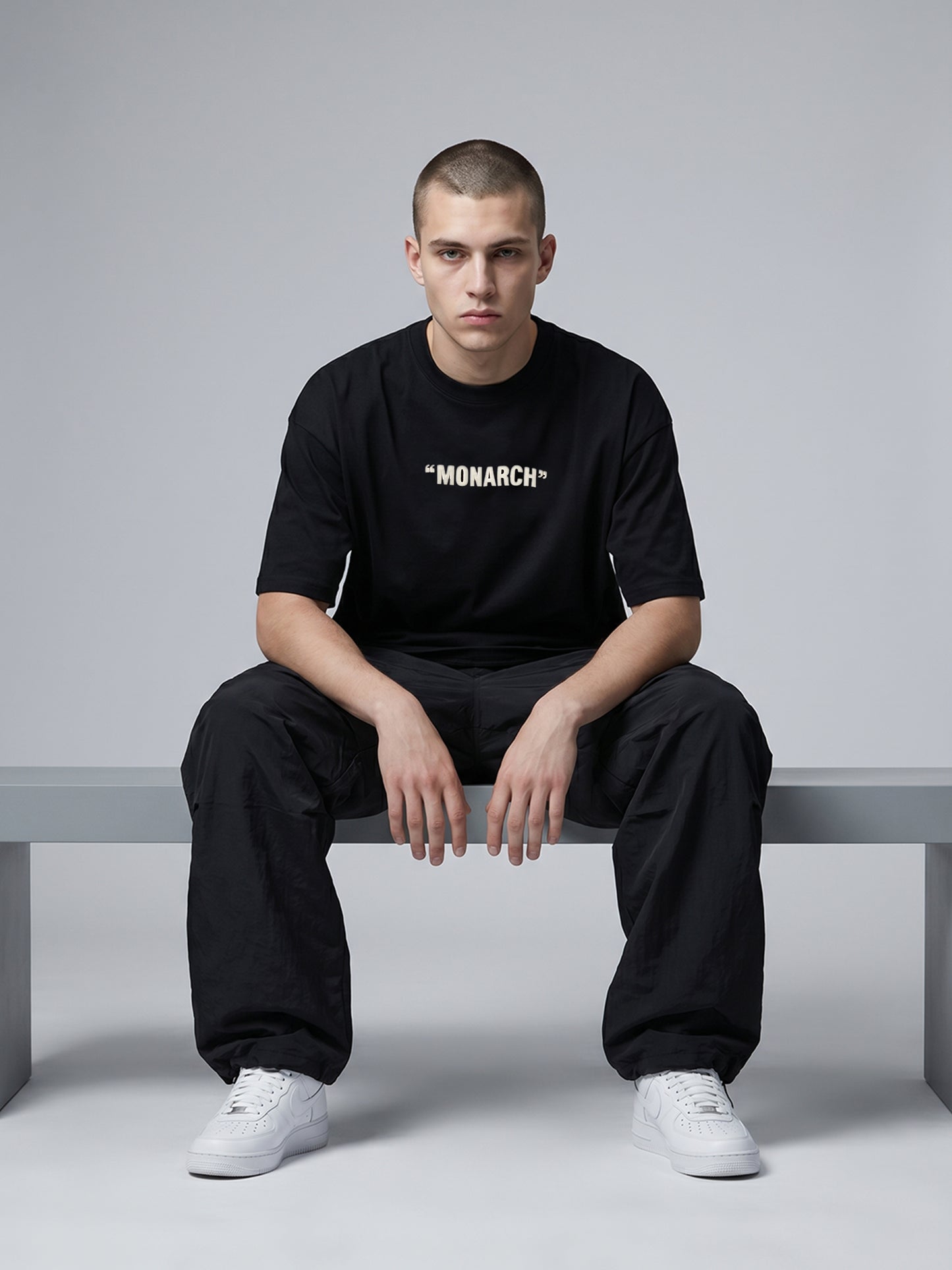 Person wearing a black 'MONARCH' t-shirt sitting on a bench against a gray background