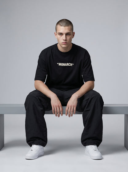 Person wearing a black 'MONARCH' t-shirt sitting on a bench against a gray background