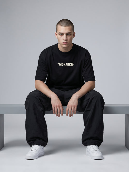 Person wearing a black 'MONARCH' t-shirt sitting on a bench against a gray background