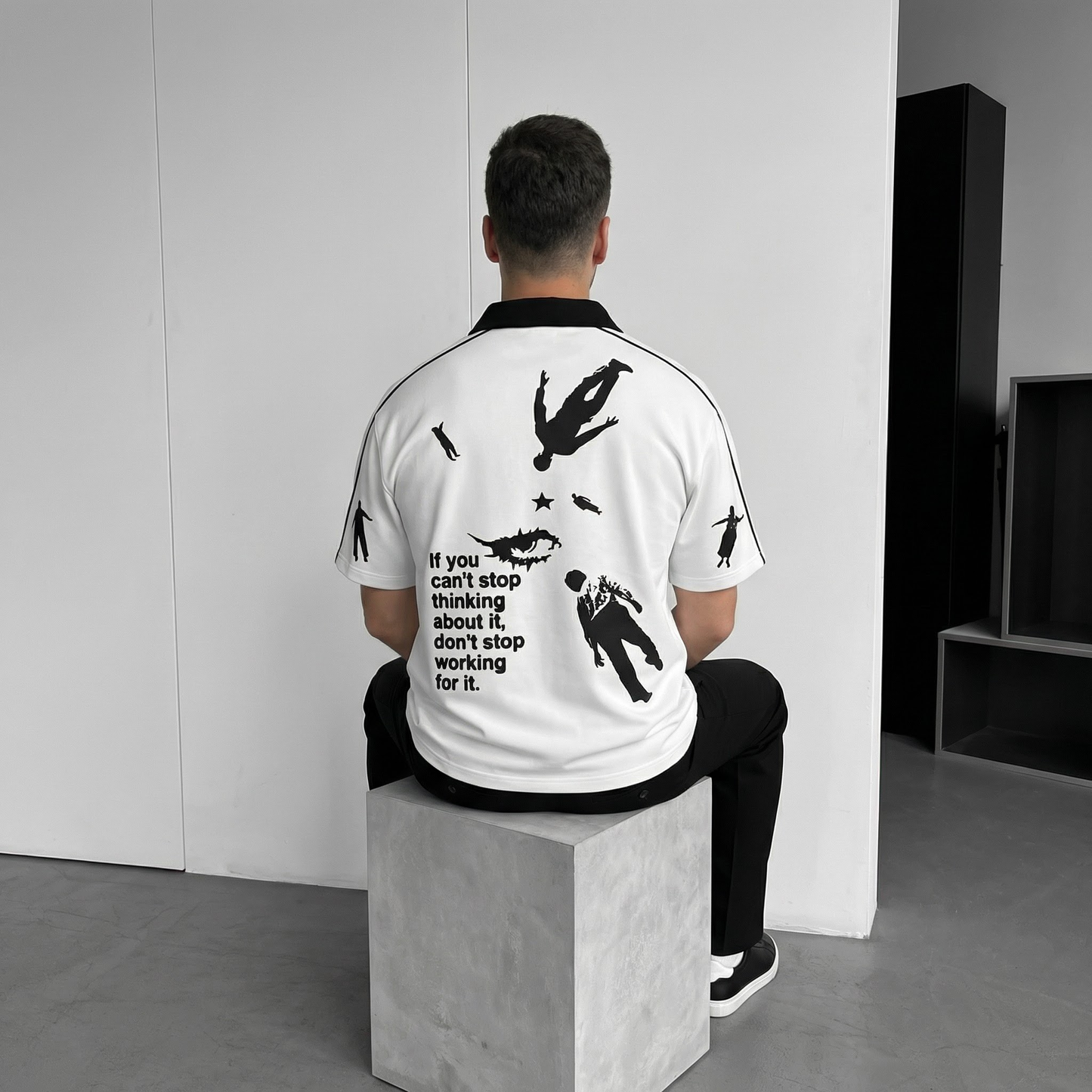 Person wearing a white polo t-shirt with black graphics and text, sitting on a block in a minimalistic setting.
