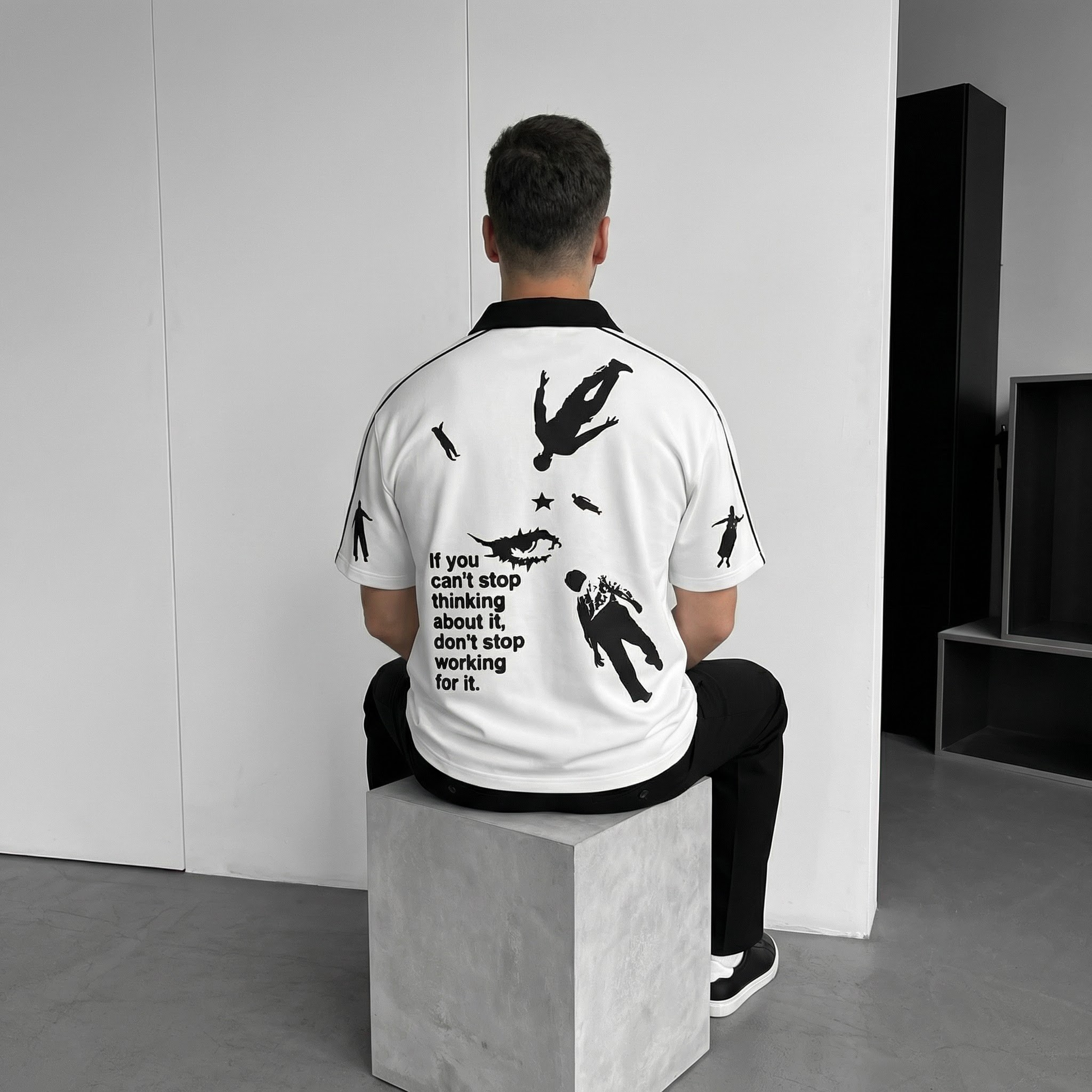 Person wearing a white polo t-shirt with black graphics and text, sitting on a block in a minimalistic setting.