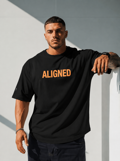 A man wearing a black crewneck t-shirt with the word 'ALIGNED' printed in orange across the chest, leaning against a light gray wall.