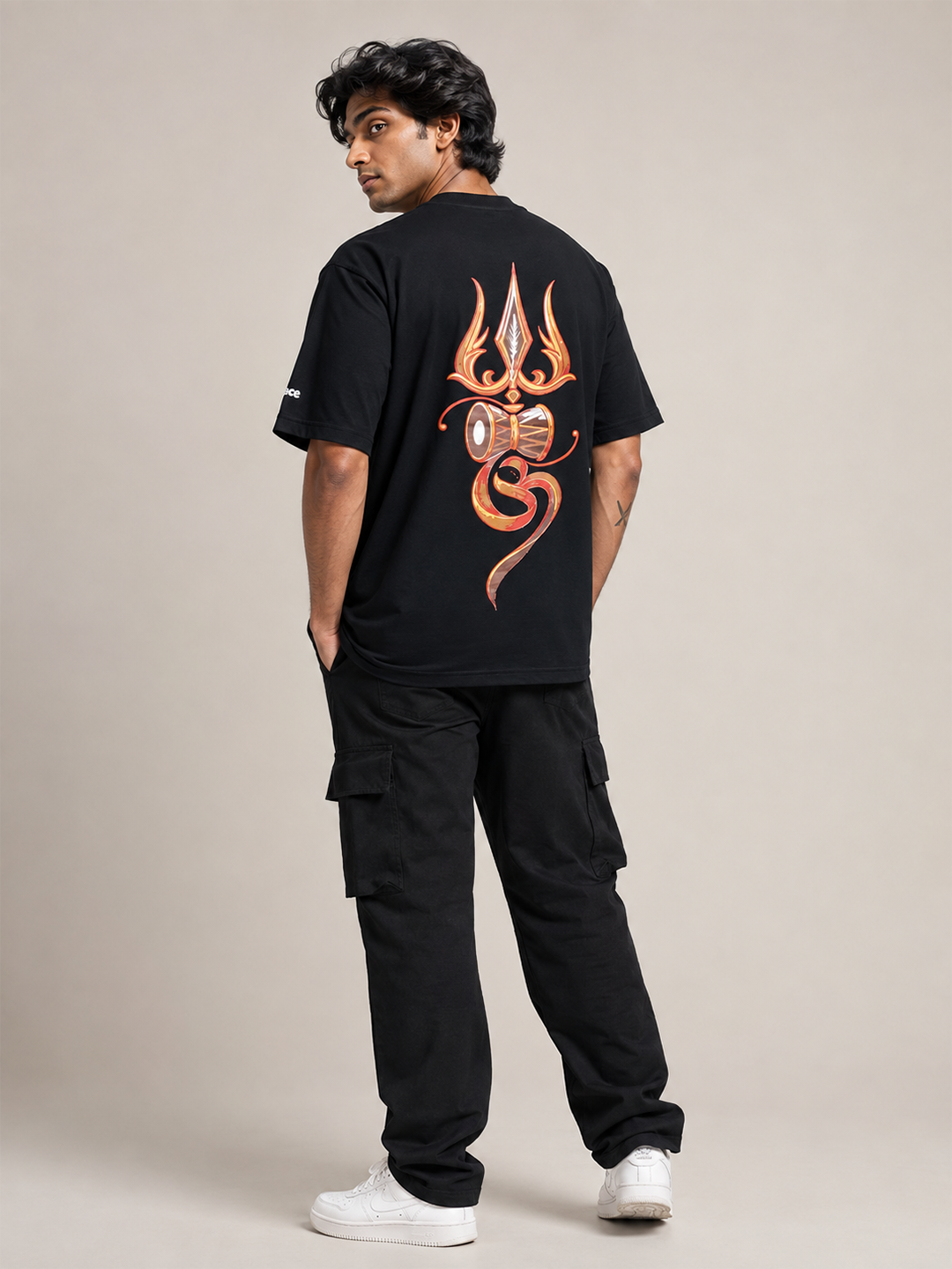 A man wearing a black t-shirt with a large orange and gold trident graphic on the back, paired with black cargo pants and white sneakers, photographed from behind against a neutral beige studio background.