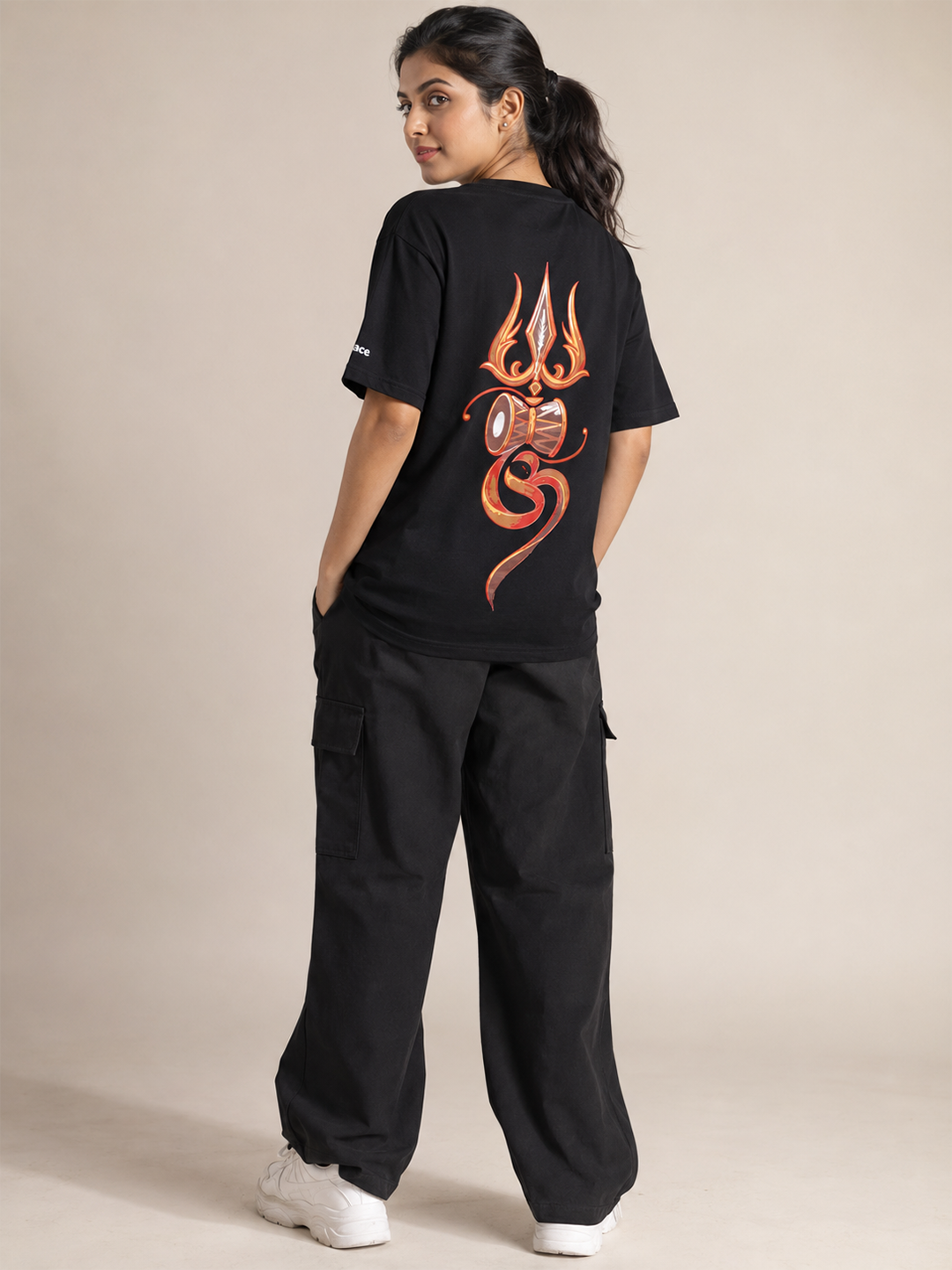 A woman modeling a black t-shirt from the back, featuring a large orange and red flame-like graphic print, paired with black cargo pants and white sneakers, set against a plain beige studio background.