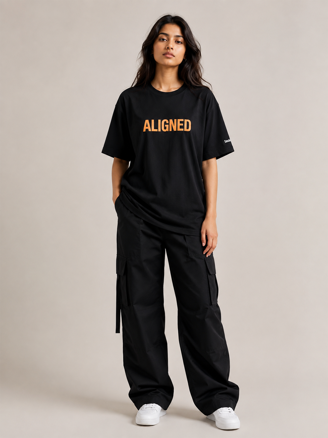 A woman modeling a black oversized T-shirt with the word 'ALIGNED' printed in orange across the chest, paired with black cargo pants and white sneakers, against a plain beige studio background.