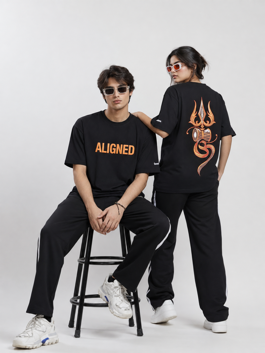 Two models wearing matching black streetwear outfits with orange graphic details, including one seated model showing the front 'ALIGNED' text on the chest and one standing model showing a large orange graphic on the back, set against a plain light gray studio background.