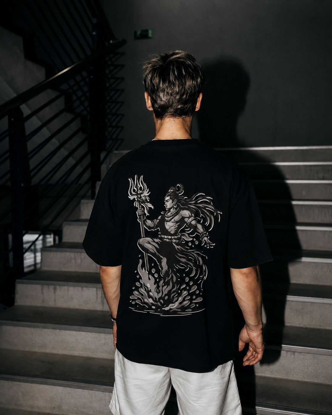 A man viewed from behind wearing a black t-shirt with a large white graphic of on the back, standing on concrete stairs in a dark indoor setting.