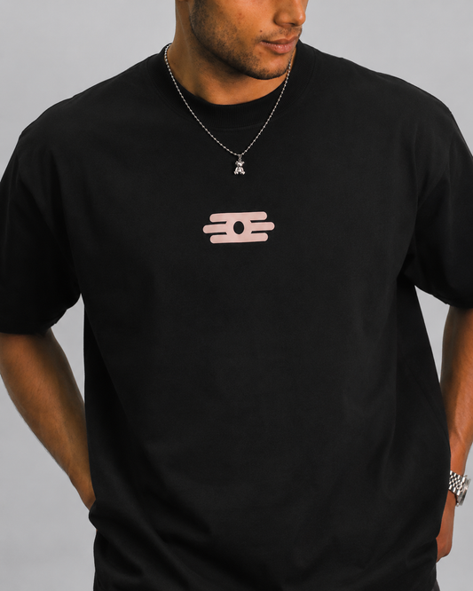 A man wearing a black crewneck t-shirt with a small light pink graphic logo on the center chest, paired with a silver chain necklace.