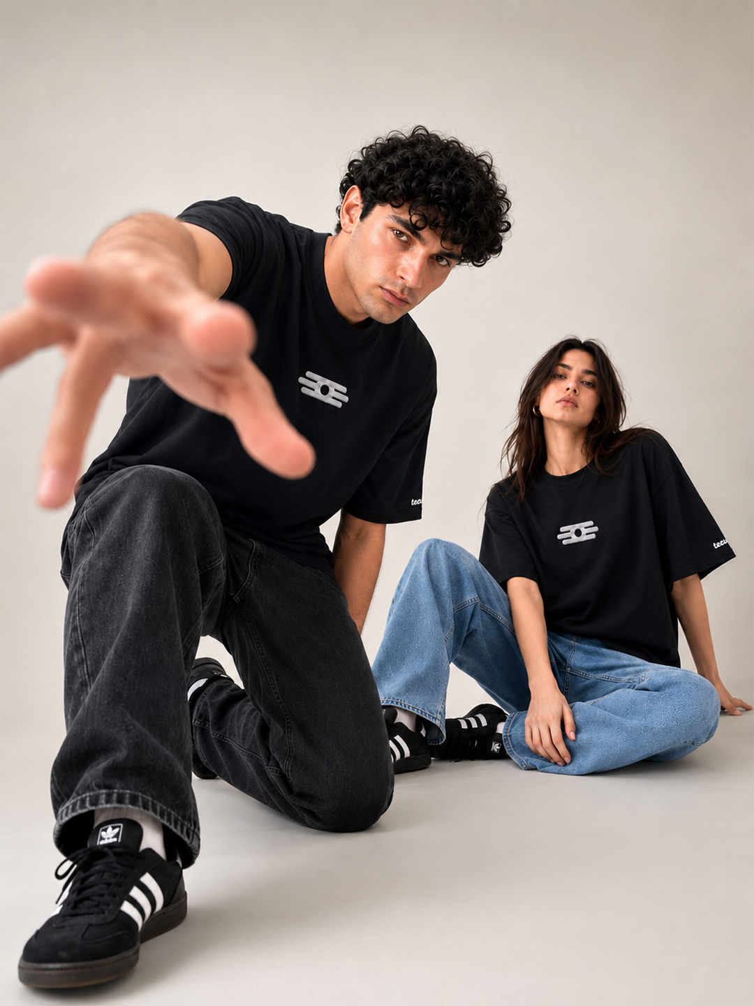 Two models wearing matching black graphic t-shirts and black sneakers with white stripes, posed in a studio setting against a neutral light gray background.