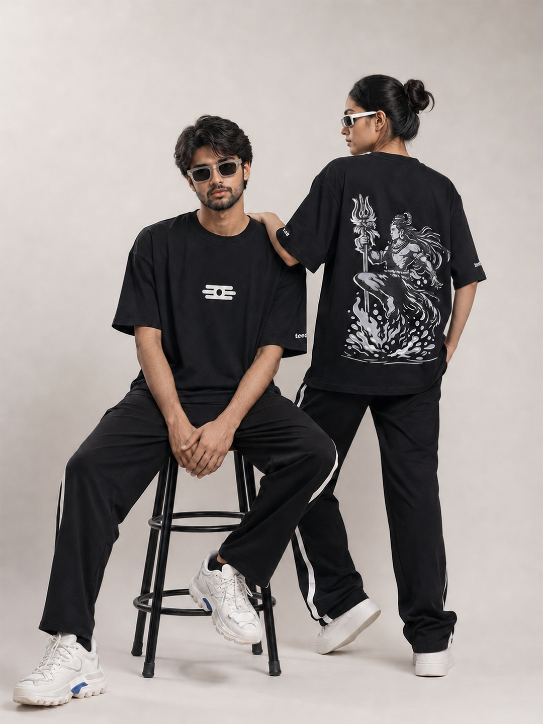 Two models wearing matching black streetwearoversized t-shirt with white graphics, posed on a light beige studio background. One model sits on a black metal stool showing the front of the black t-shirt with a white logo, while the other stands showing the back of the black t-shirt with a large white graphic print.