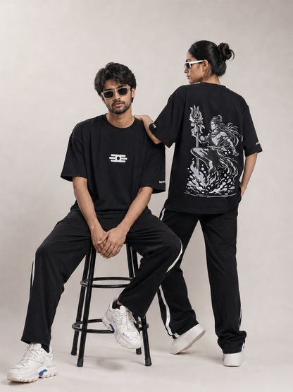Two models wearing matching black streetwearoversized t-shirt with white graphics, posed on a light beige studio background. One model sits on a black metal stool showing the front of the black t-shirt with a white logo, while the other stands showing the back of the black t-shirt with a large white graphic print.