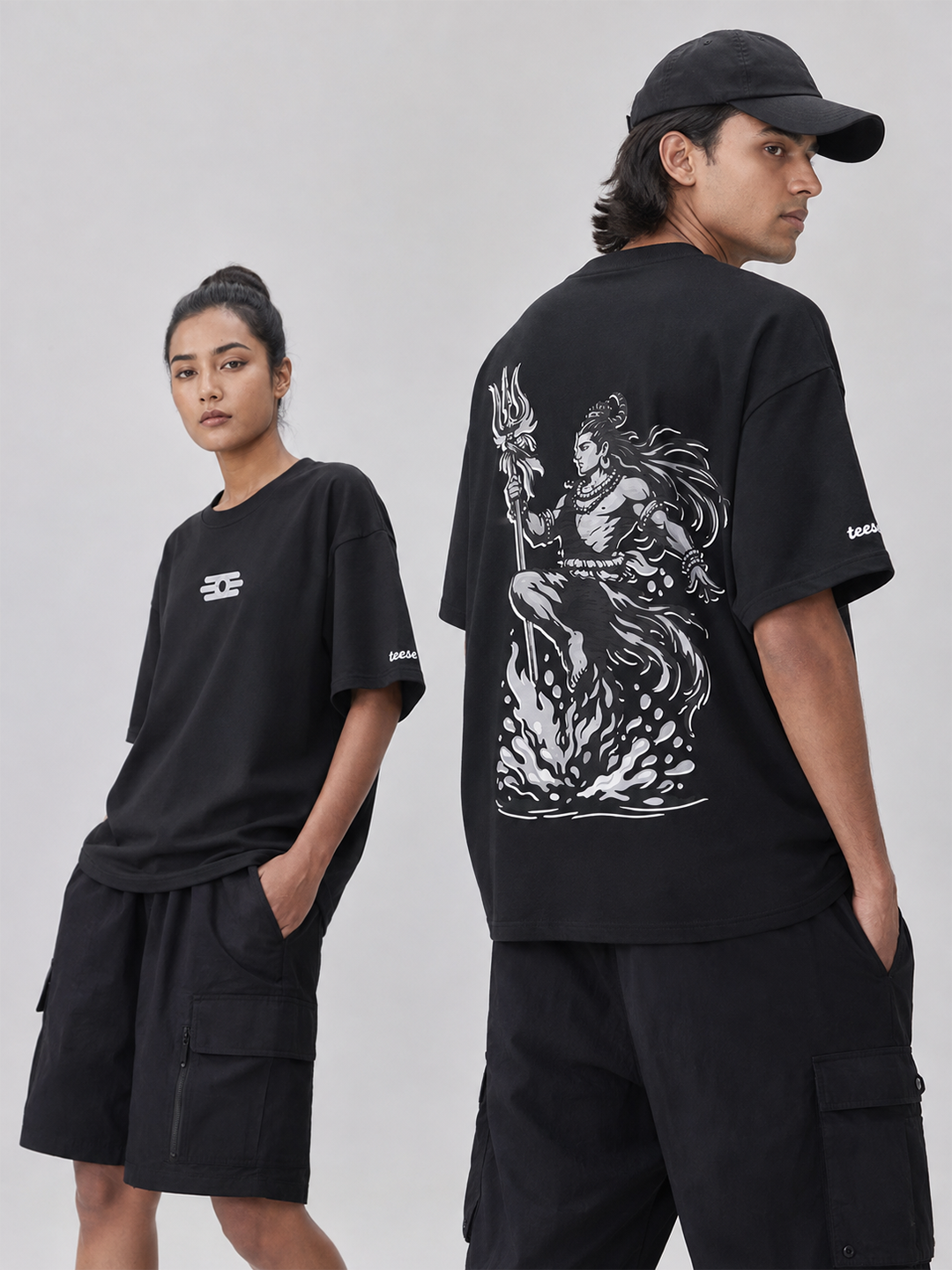Two models wearing black oversized graphic T-shirts and black cargo shorts against a light gray studio background. The male model shows the back of the shirt with a large white line-art graphic, while the female model shows the front with a small white chest logo.